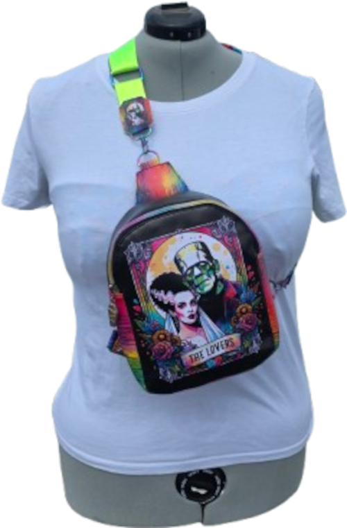 A black crossbody bag with a colorful 'The Lovebirds' graphic print, displayed on a white t-shirt dress mannequin. The bag features a vibrant, multicolored illustration of Frankenstein and Bratz, with a bright neon green strap.