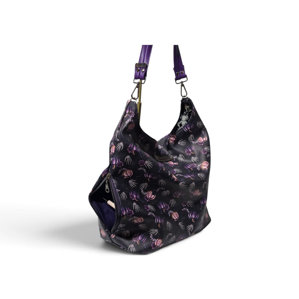 A black slouchy shoulder bag with a purple and pumpkin pattern, featuring purple straps shown on a white background.
