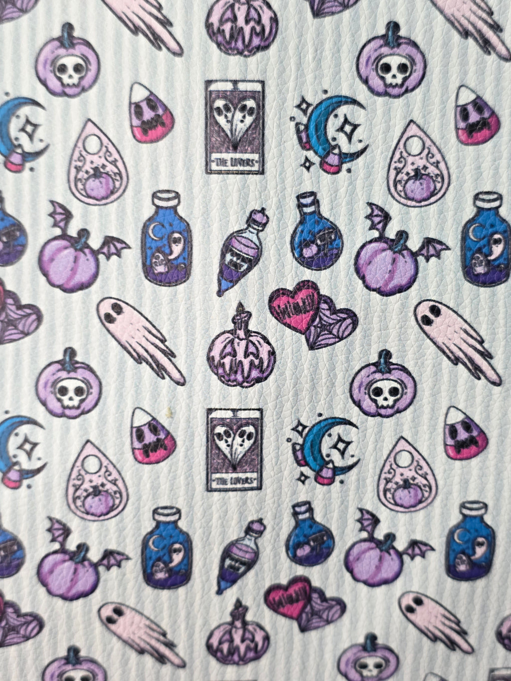 A close-up of a white textured fabric featuring a repeating pattern of whimsical, hand-drawn Halloween-themed illustrations in purple, pink, and blue, including pumpkins, skulls, ghosts, and tarot cards.