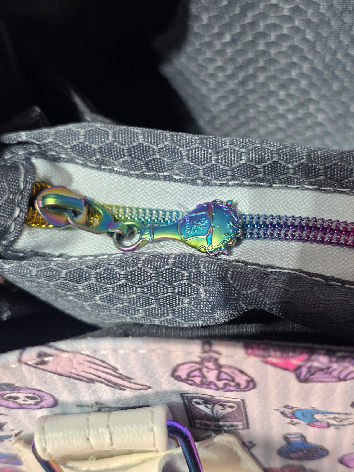 Close-up of a metallic zipper with an iridescent rainbow-colored pull tab and matching zipper tape, set against a gray patterned fabric background.