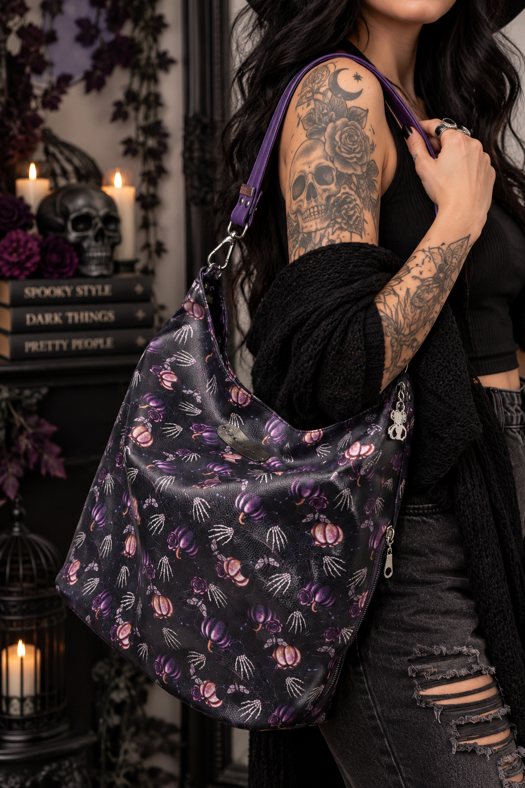 A woman with tattooed arms carries a large black shoulder bag decorated with a purple and pink gothic pumpkin and skeleton print, set against a dark, spooky-themed background with candles and books.