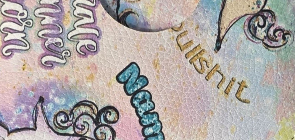 A close-up of a textured light pink surface with decorative script text in purple, gold, and blue, along with black ornamental flourishes.