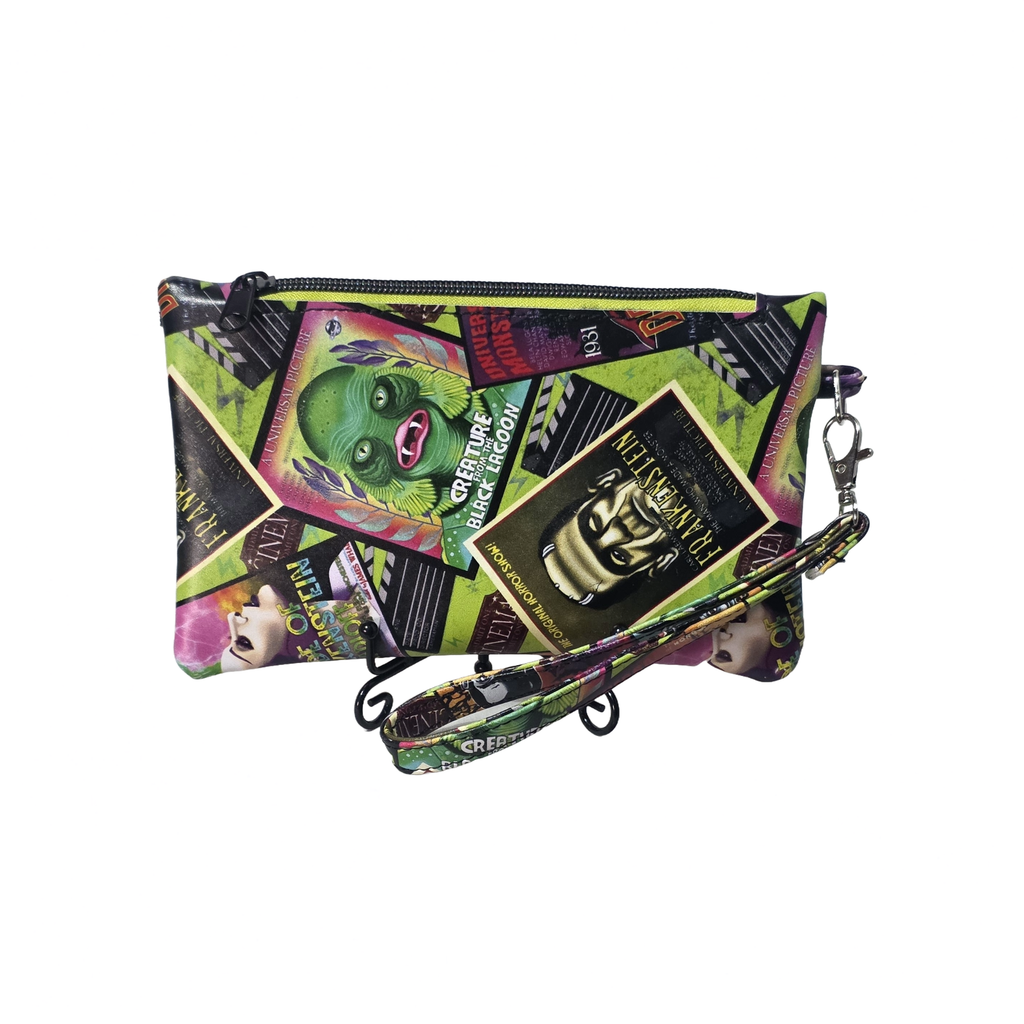 A zippered clutch bag with a vibrant collage print featuring movie posters, a green monster face, and horror-themed graphics, complete with a matching wrist strap and metal clasp.