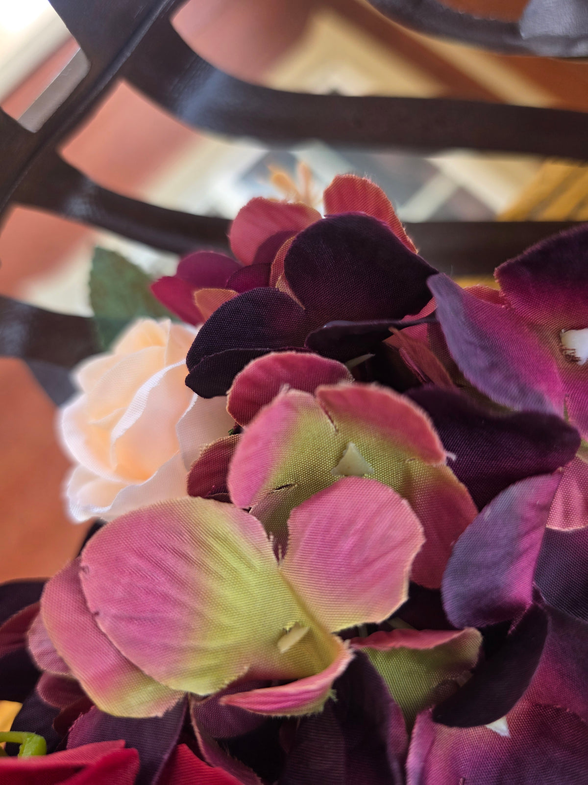 Close-up of artificial flowers, featuring dark burgundy and pink hydrangea-like blooms with pale yellow-green centers, alongside a soft peach rose, set against a blurred dark metal background.