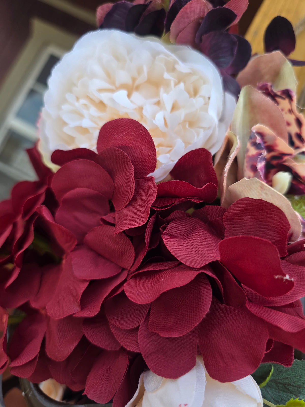 A close-up of a floral arrangement featuring deep burgundy hydrangea-like blooms, a large off-white peony, and a spotted orchid with cream, burgundy, and brown tones.