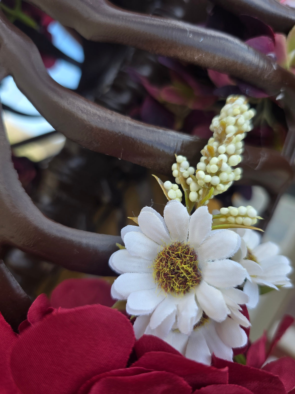 A close-up of a decorative floral arrangement featuring a prominent white daisy with a yellow-brown center, cream-colored buds, and deep red fabric petals, set against a dark brown wavy background.