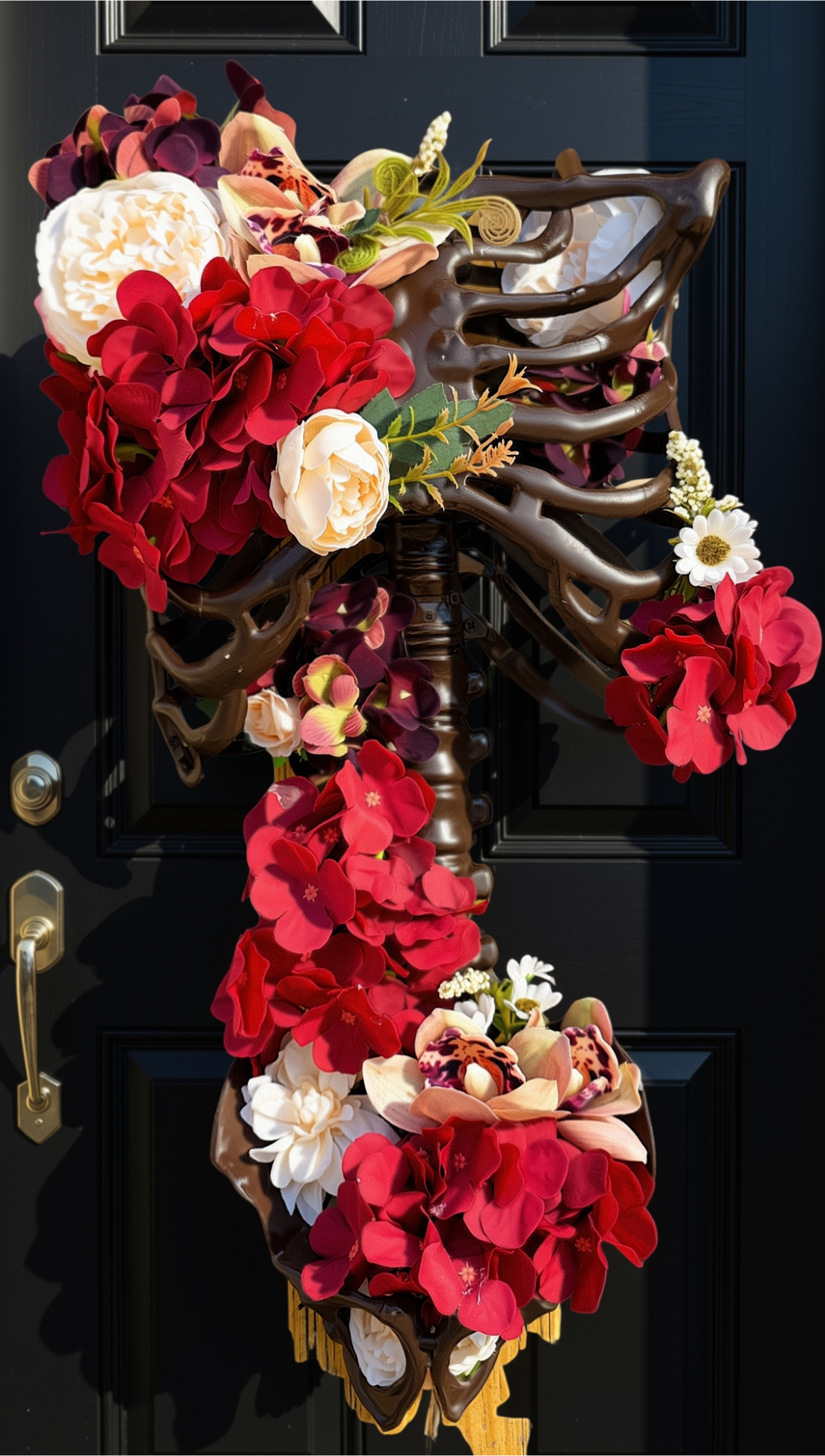 A decorative skeleton-themed door wreath hanging on a black paneled door. The wreath features a dark brown rib cage design filled with red and burgundy flowers, cream roses, small white daisies, and green foliage accents.